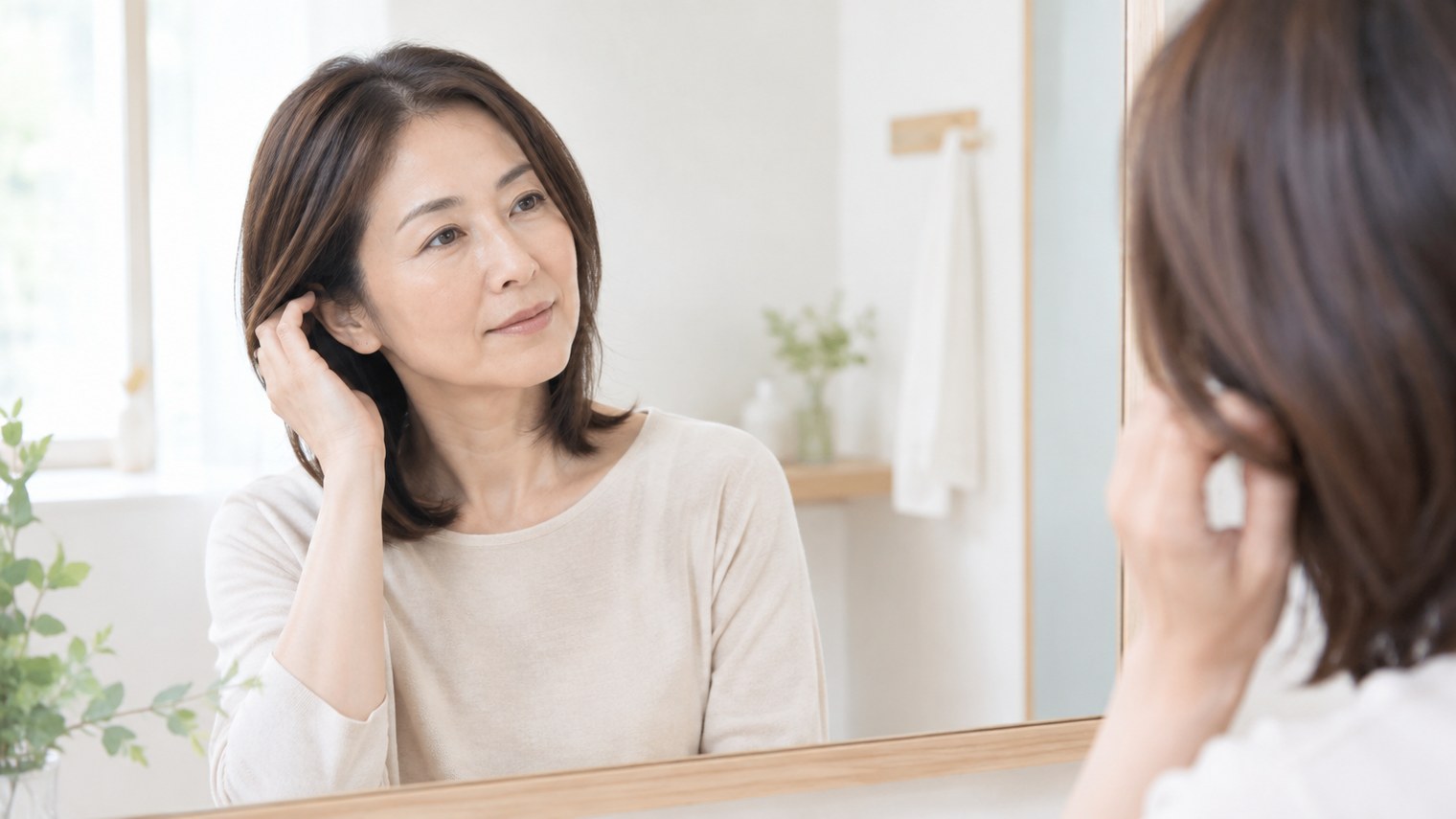 明るい洗面所で、鏡の前で髪を耳にかけながら、落ち着いた表情でいる女性のイメージ写真。