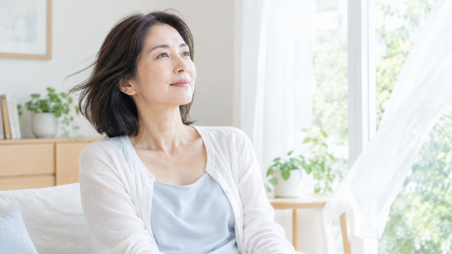明るい窓辺で、カーディガンの前を開けて風に当たり、一息ついている50代前後の女性のイメージ写真。